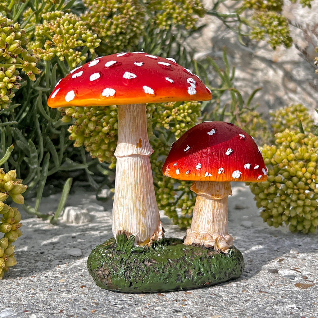 Two small decorative toadfoot mushroom garden figurines with red caps, white spots, and beige stems, placed on a mossy base among green plants and stone.