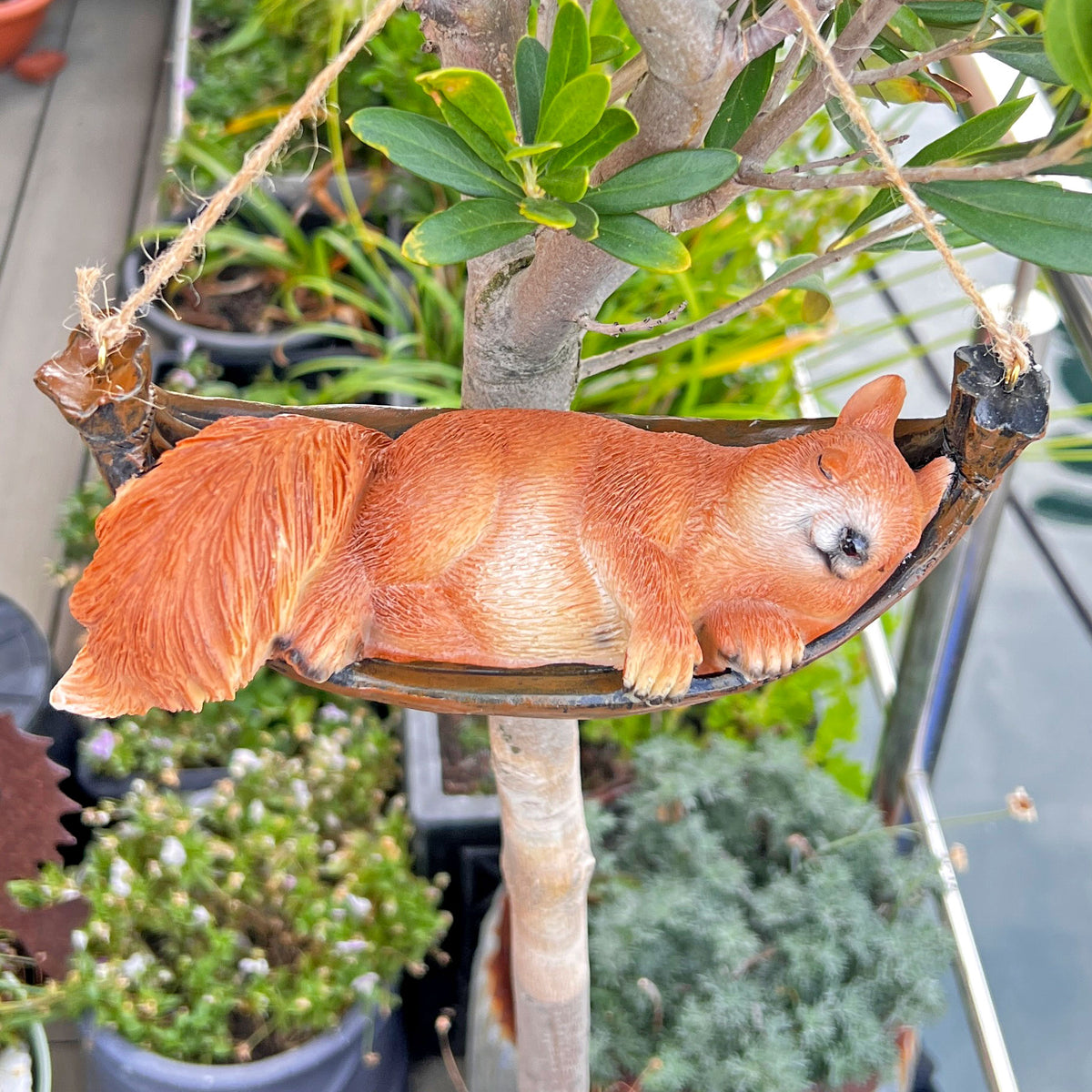A sleeping squirrel garden ornament resting in a hammock, hanging from a tree in a potted garden setting.