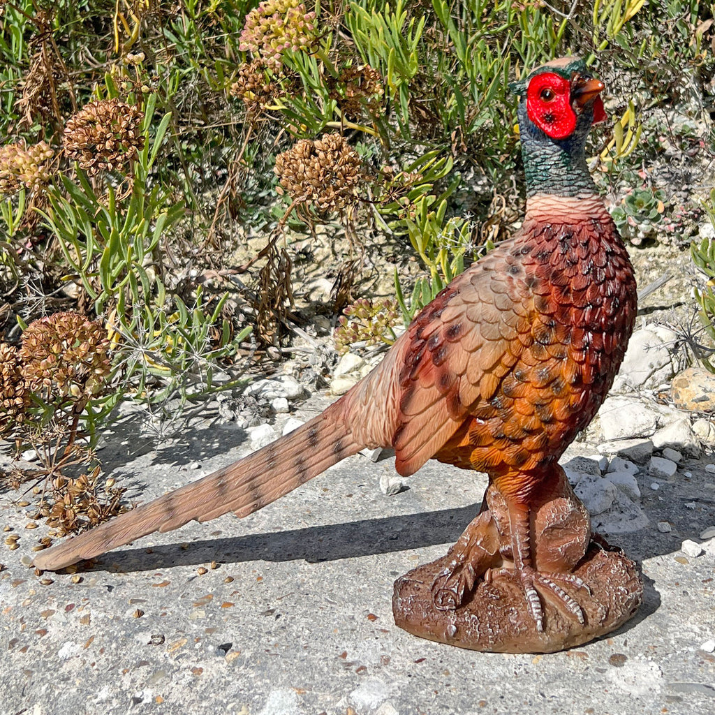 A realistic pheasant bird statue or decorative figure standing on a textured brown base outdoors on a stone surface, with green plants and dry flowers in the background.