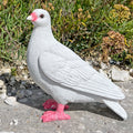 A realistic white garden pigeon statue with pink beak and feet, standing on a stone surface outdoors with plants in the background.